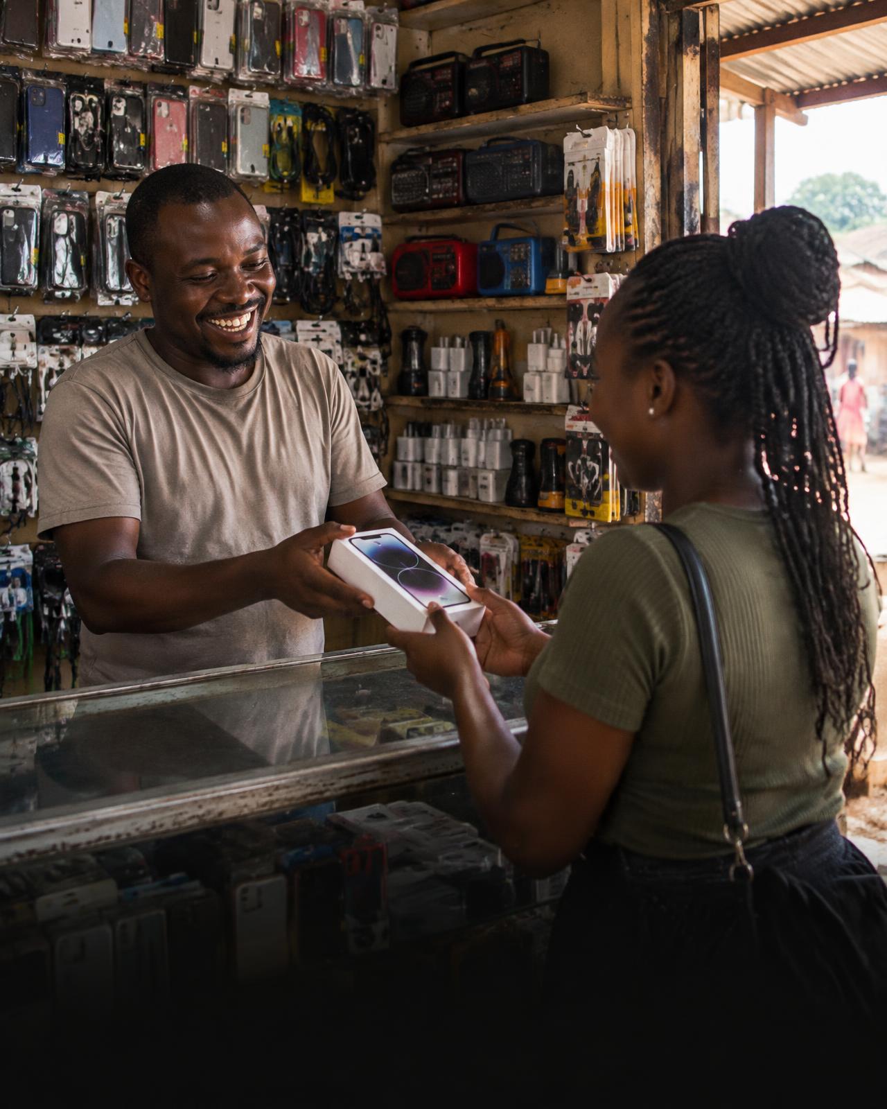 A shopkeeper handing a boxed phone to a smiling customer in a Kampala electronics shop